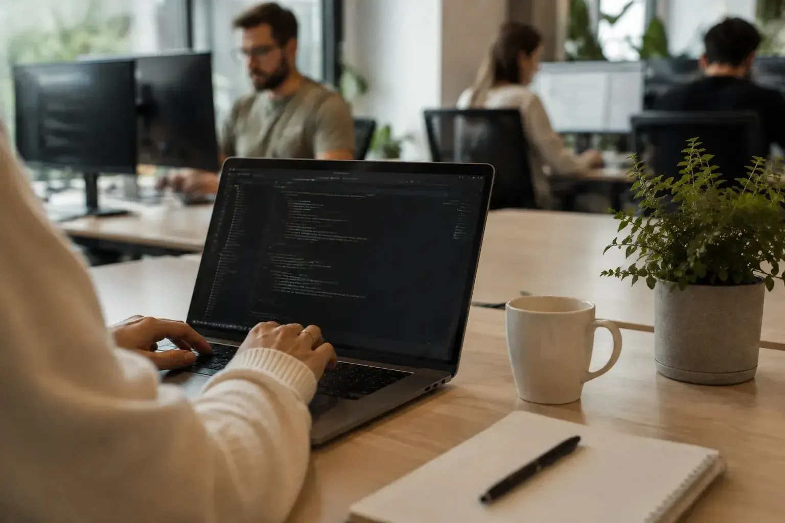 A person writes code on a laptop in the foreground while colleagues work in the background of an open-plan office.