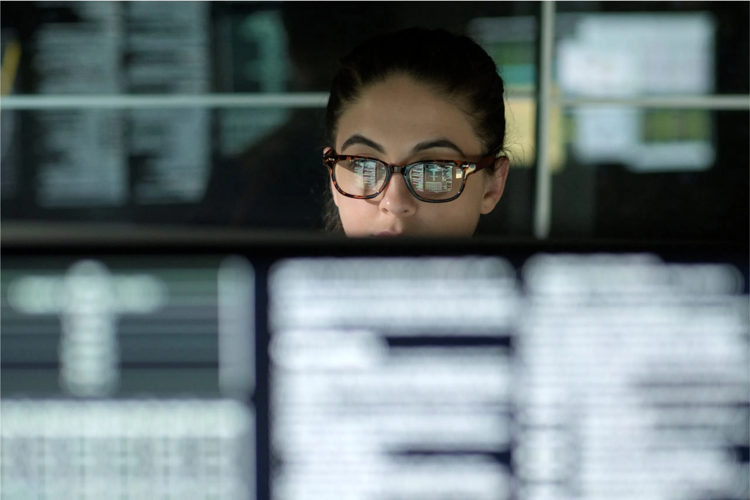 A woman with glasses, lines of code reflected in her lenses, looks attentively over a monitor.