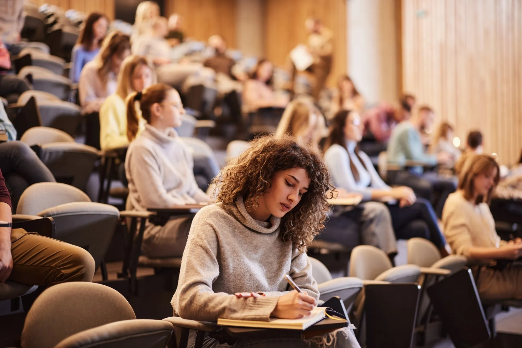 A student takes notes in her notebook in a tiered lecture hall, with other students seated behind her.