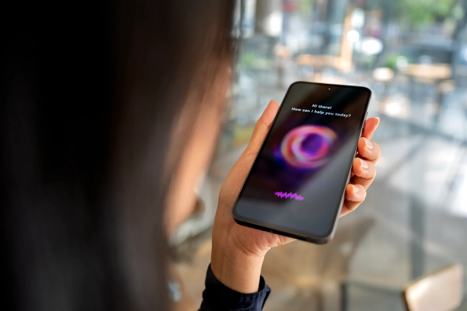 A woman holds a mobile phone whose screen shows a voice assistant asking how it can help.