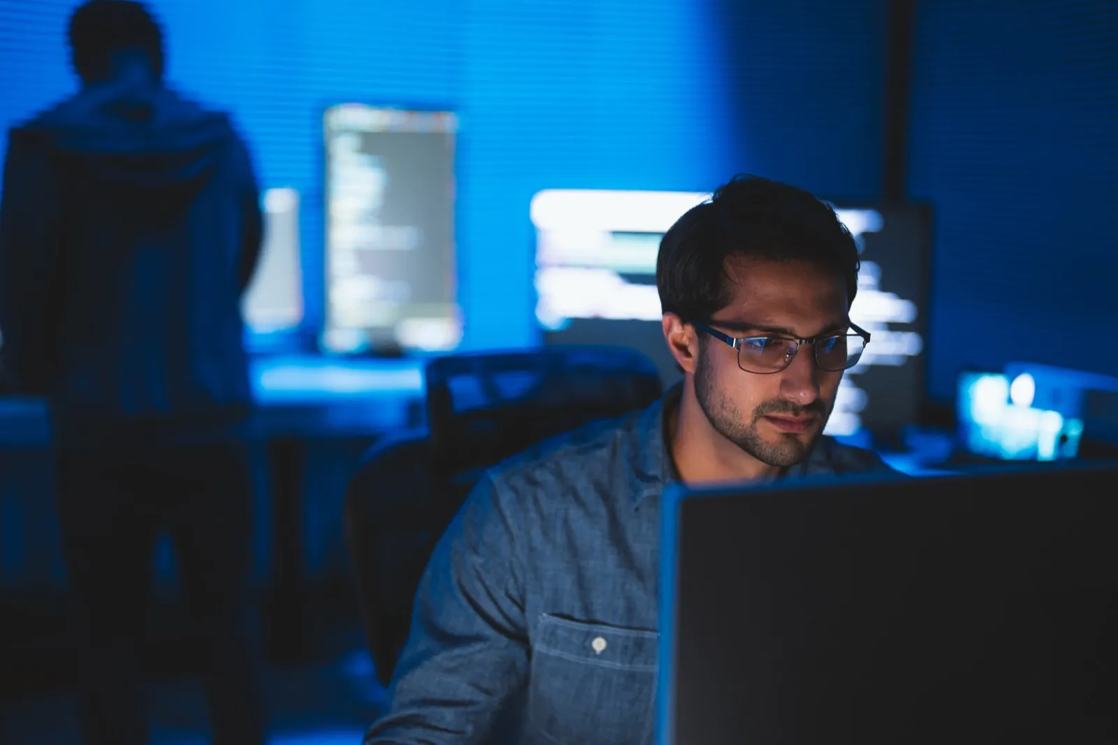 A man wearing glasses works at a monitor in a dimly lit room bathed in blue, with a colleague in the background.
