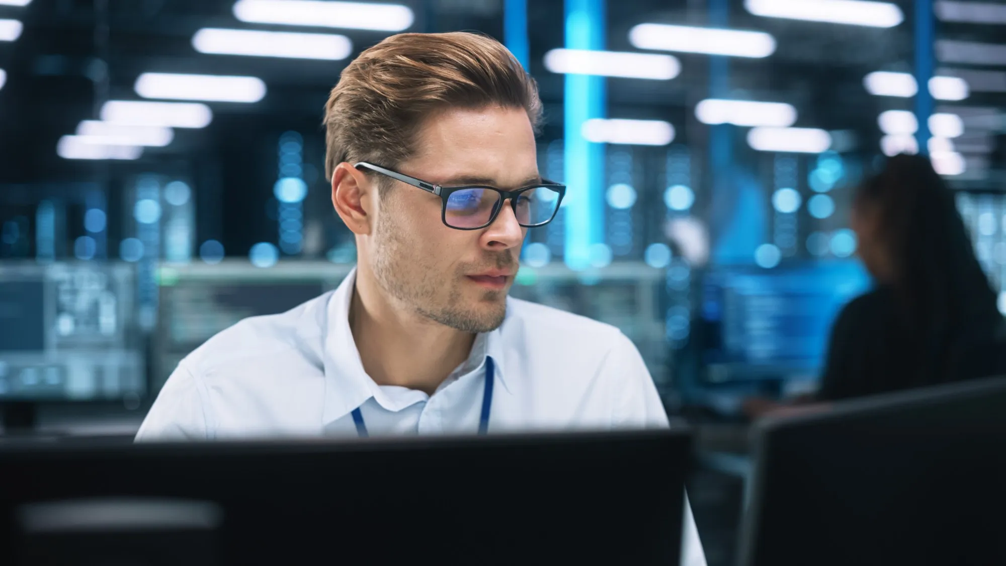 A man in glasses works intently at his monitor in a technical room with blue lights in the background.