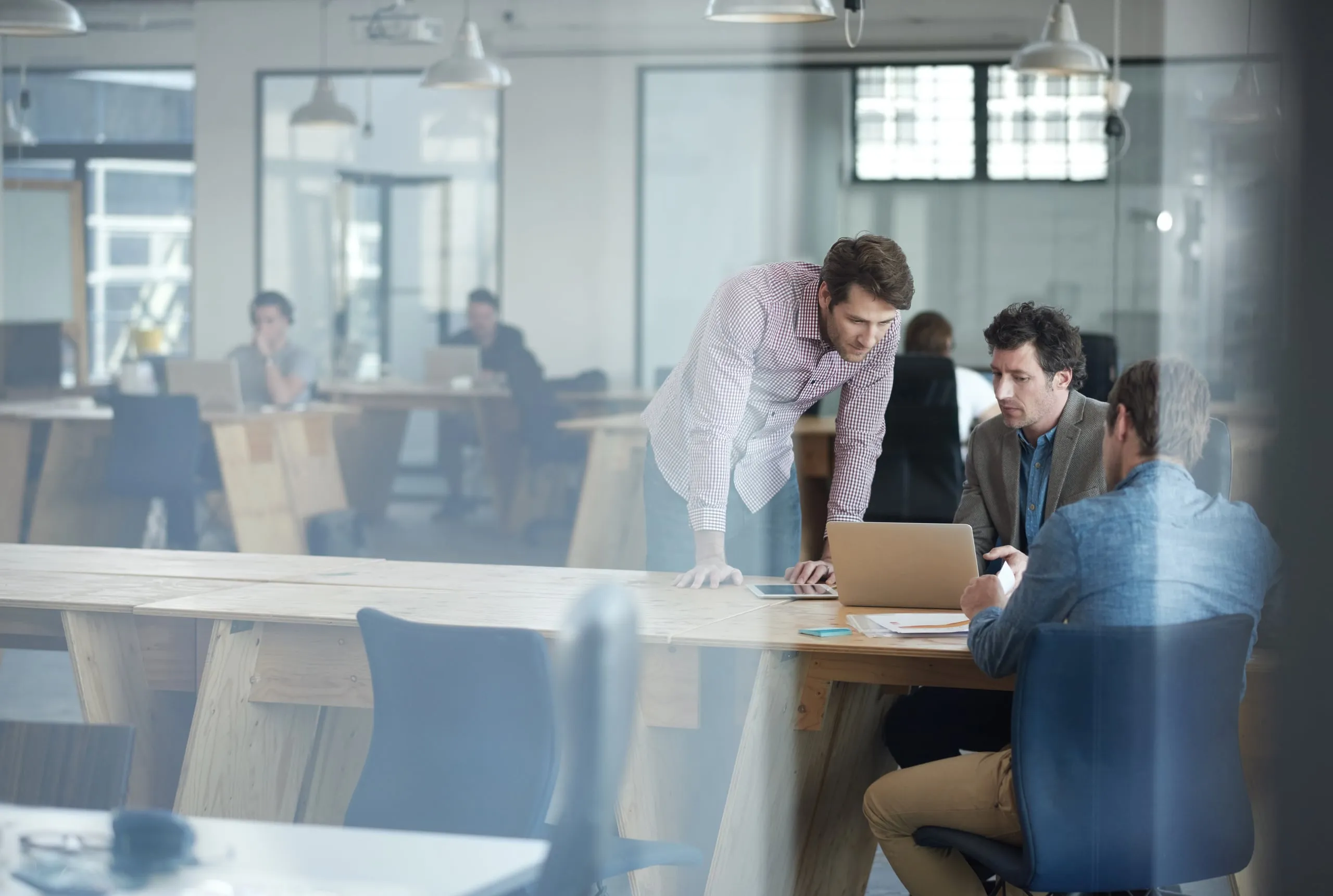 Tres compañeros revisan juntos un portátil en una mesa de trabajo de una oficina diáfana.