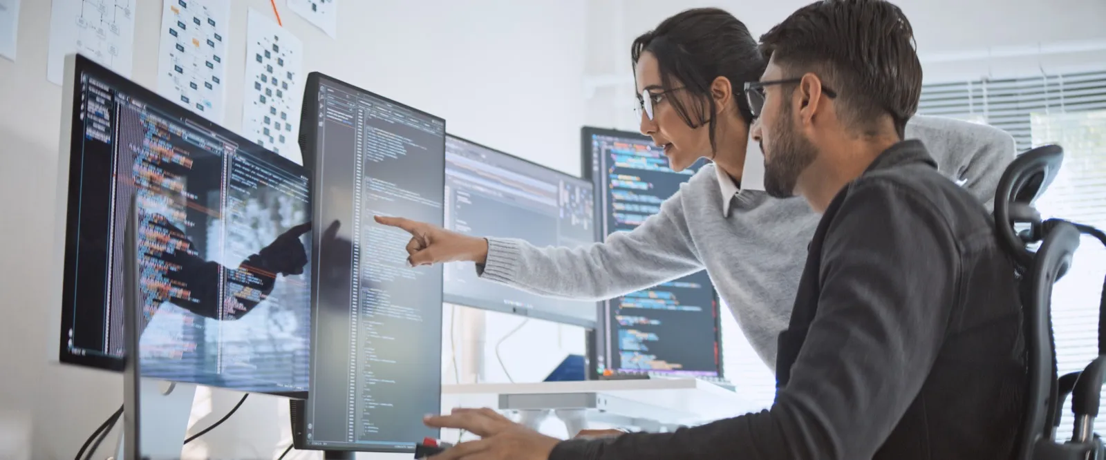 A woman points at code on a monitor while a colleague sitting next to her follows the explanation.