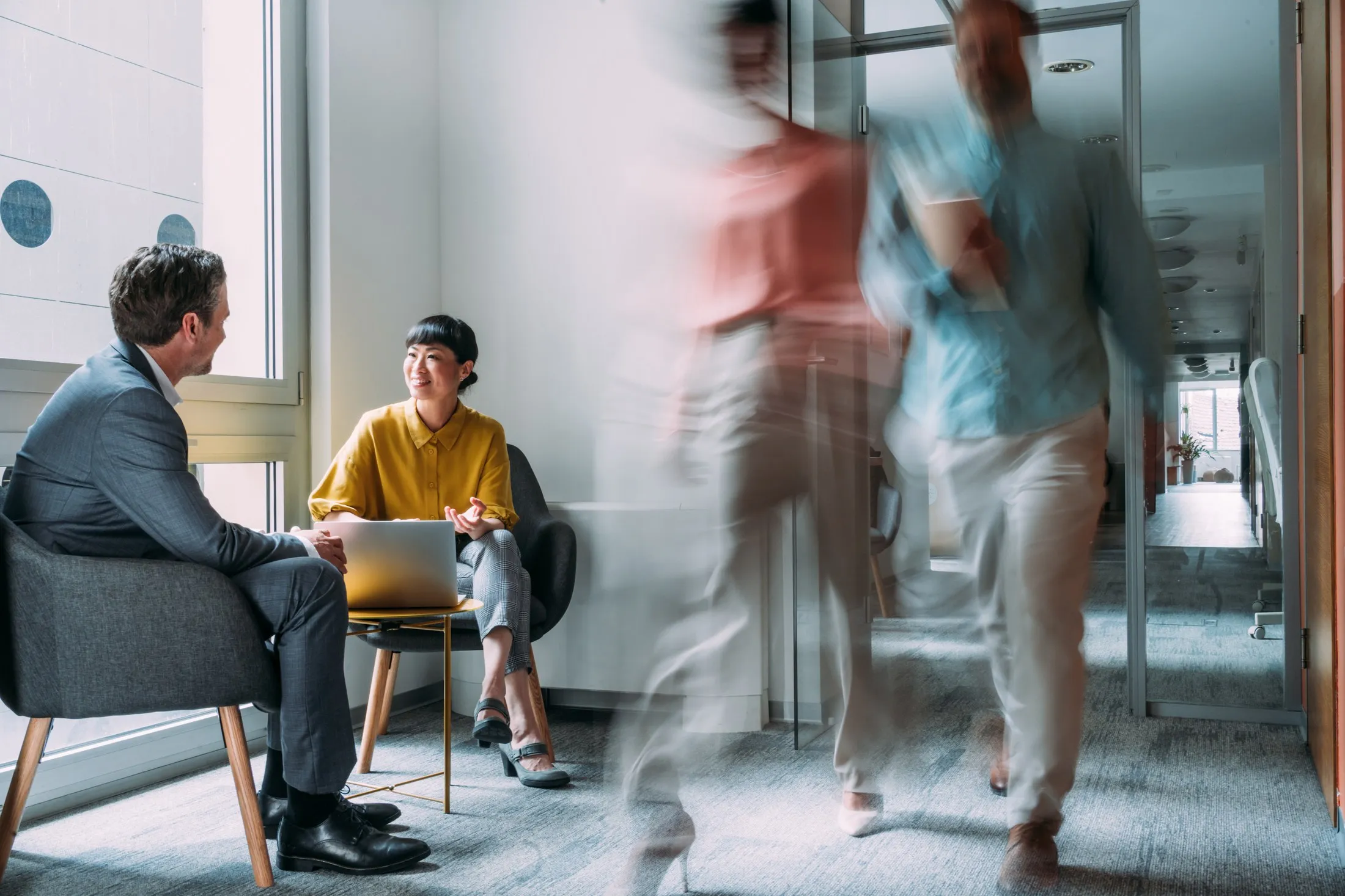 Two people talking by a window while the rest of the team moves around them.