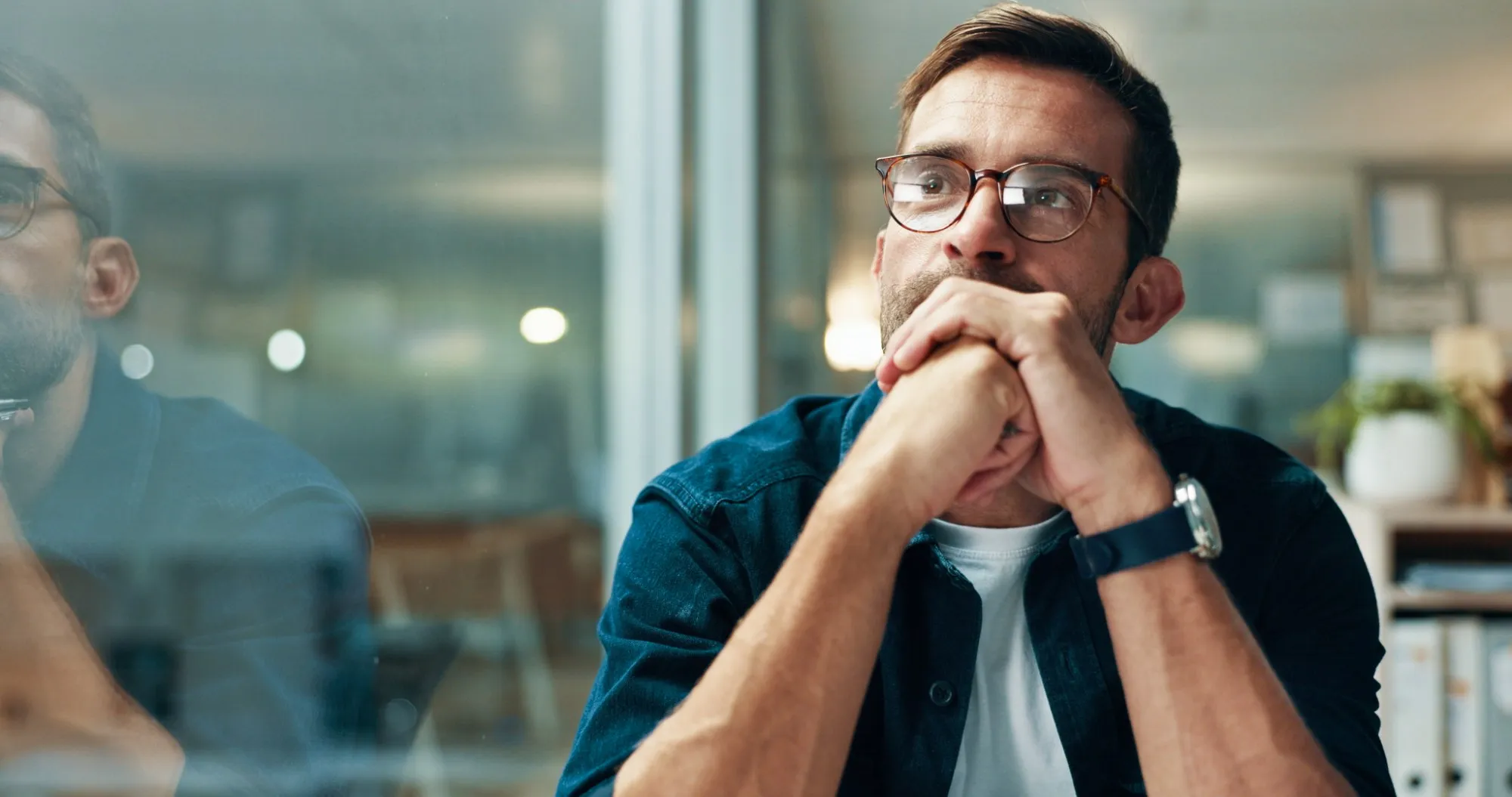 A man in glasses, hands clasped under his chin, looks thoughtfully to one side in an office.