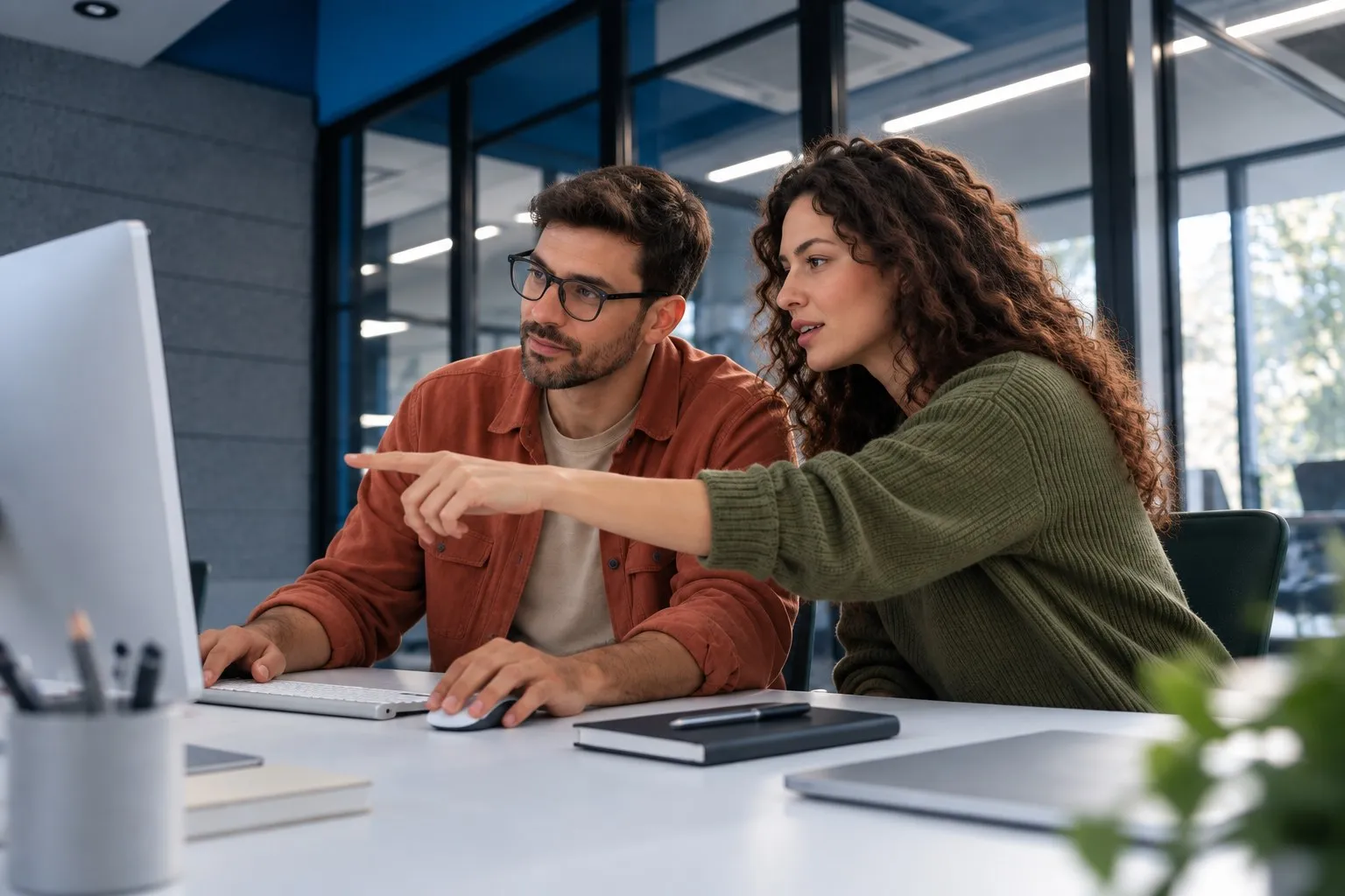 A woman points at a computer screen while a colleague in glasses listens.