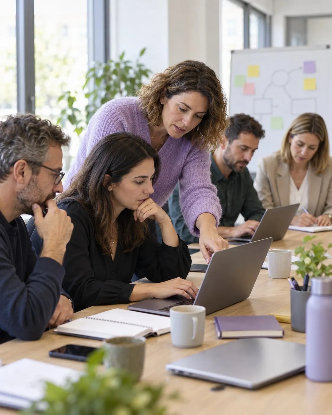 A trainer in a lilac jumper points at a participant's laptop during a workshop where several professionals work around a table.