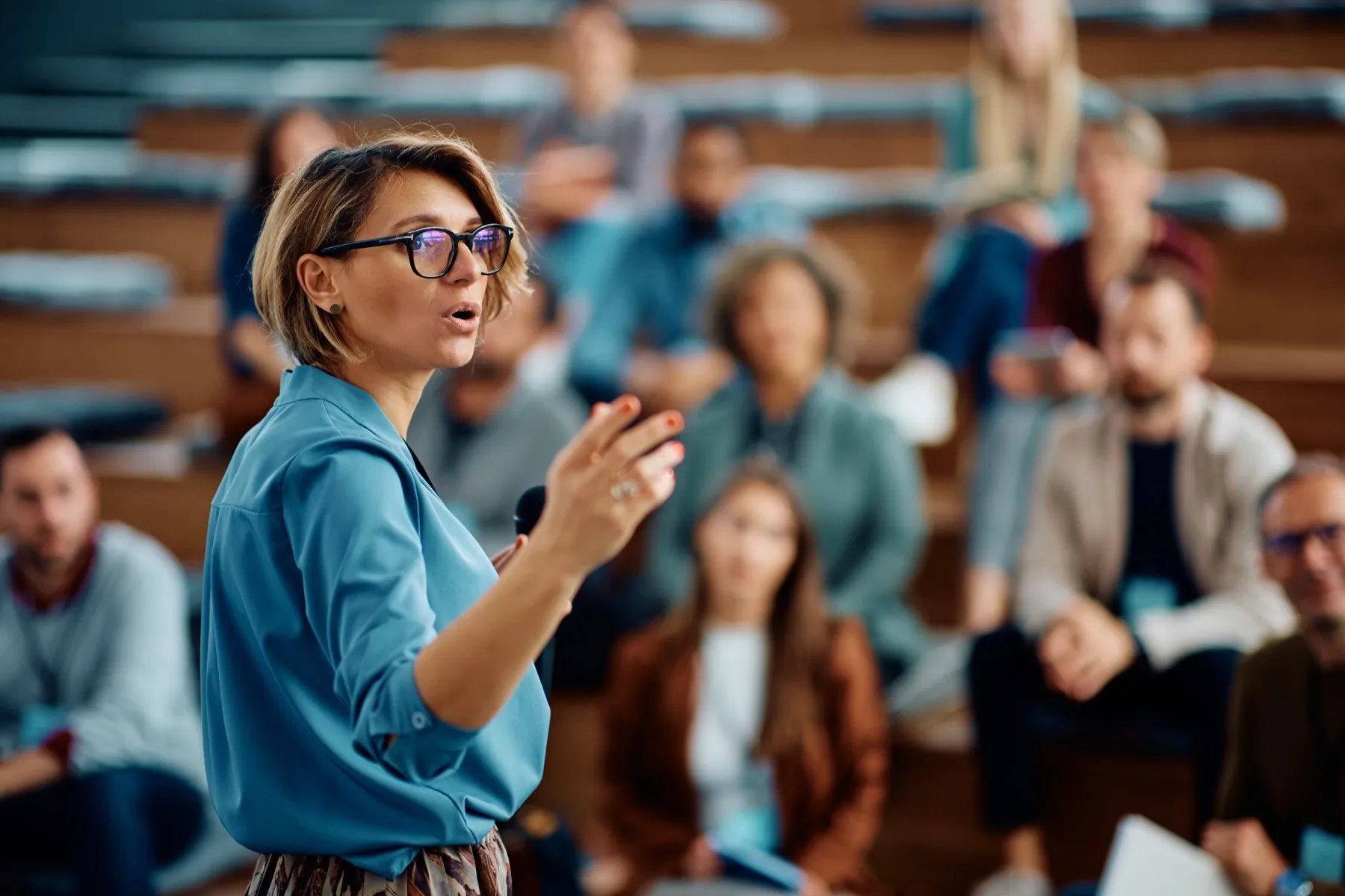 A speaker with a microphone gestures as she addresses a group seated in a tiered lecture hall.