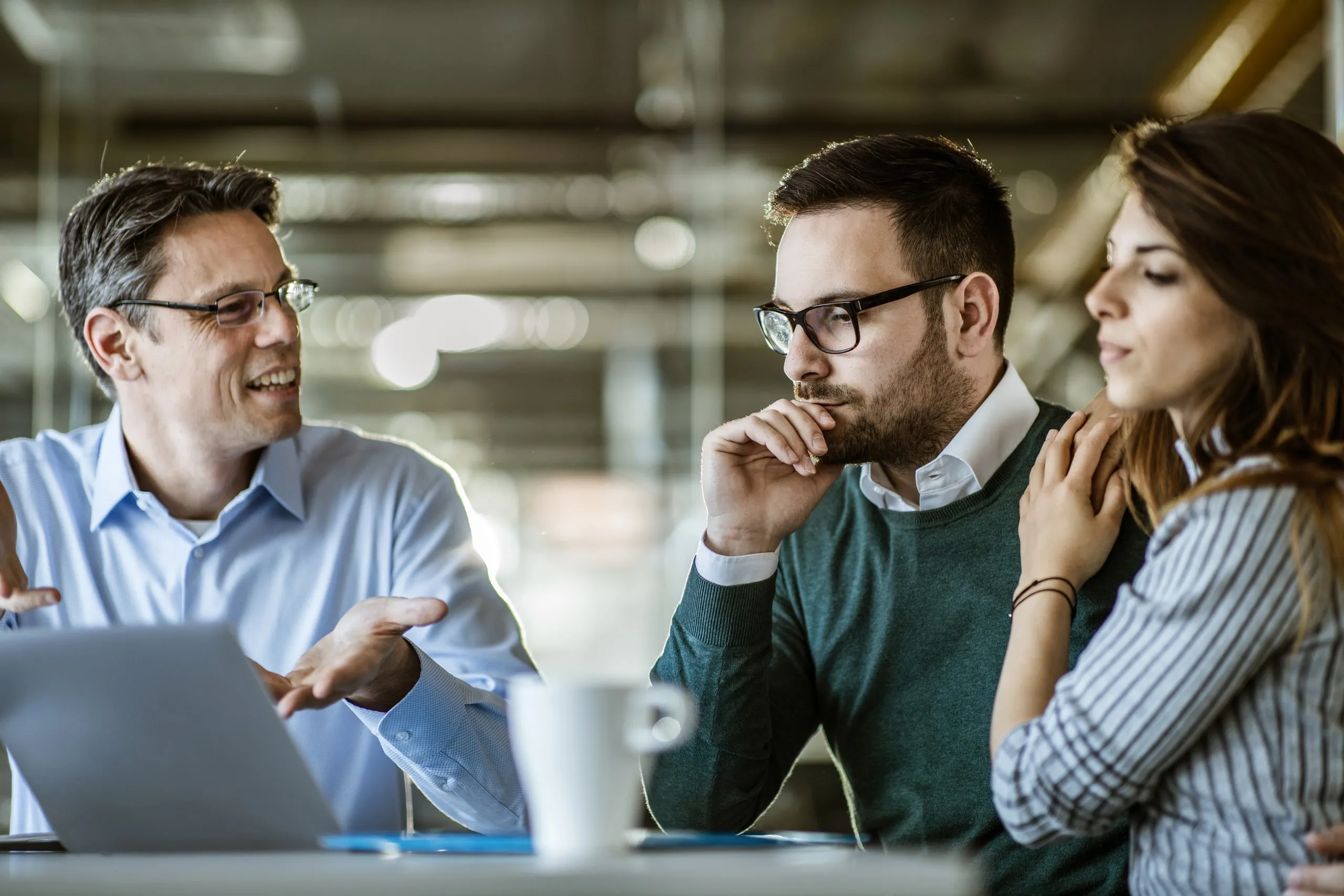 Tres personas conversan alrededor de una mesa de trabajo con un portátil delante.