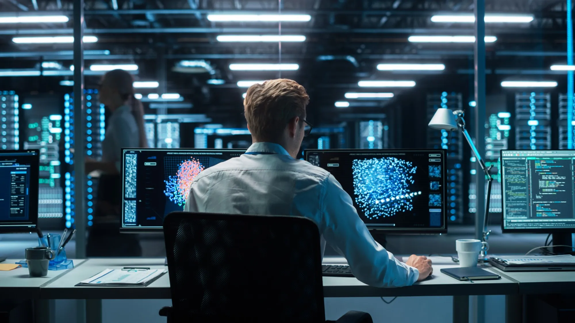A technician, seen from behind, works at several monitors showing data visualisations in a glass-walled room next to a data centre.