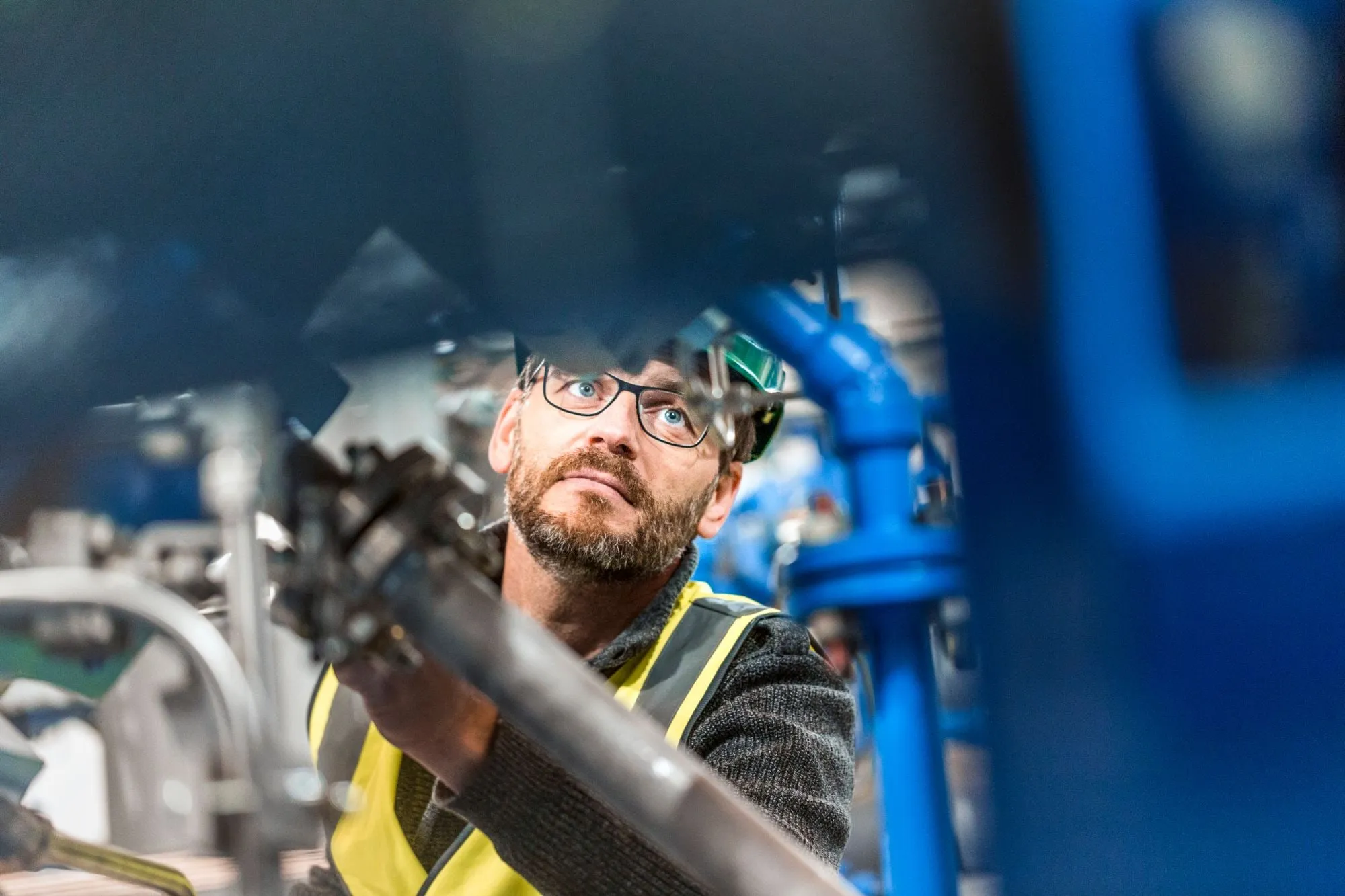 A technician in glasses and a high-visibility vest carefully inspects a machine mechanism in an industrial plant.