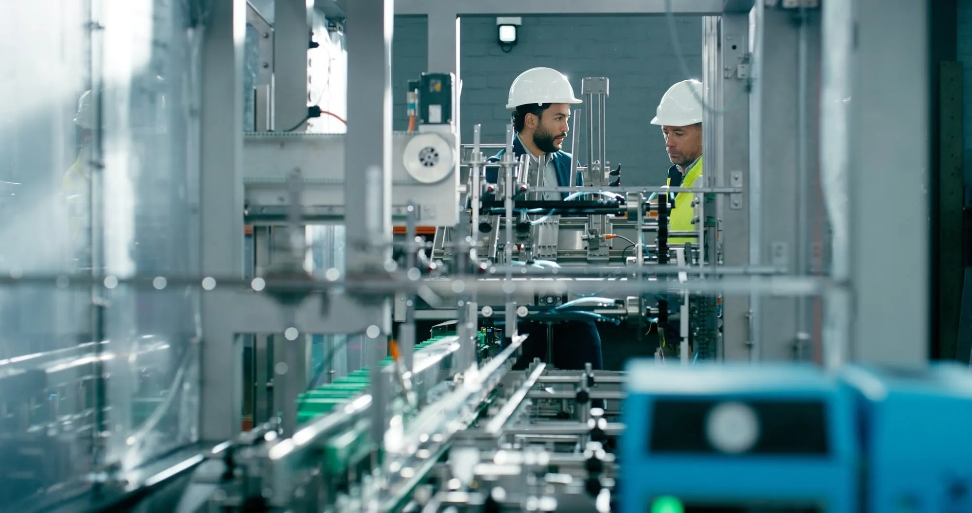 Two technicians in hard hats talk beside an automated packaging line in an industrial plant.
