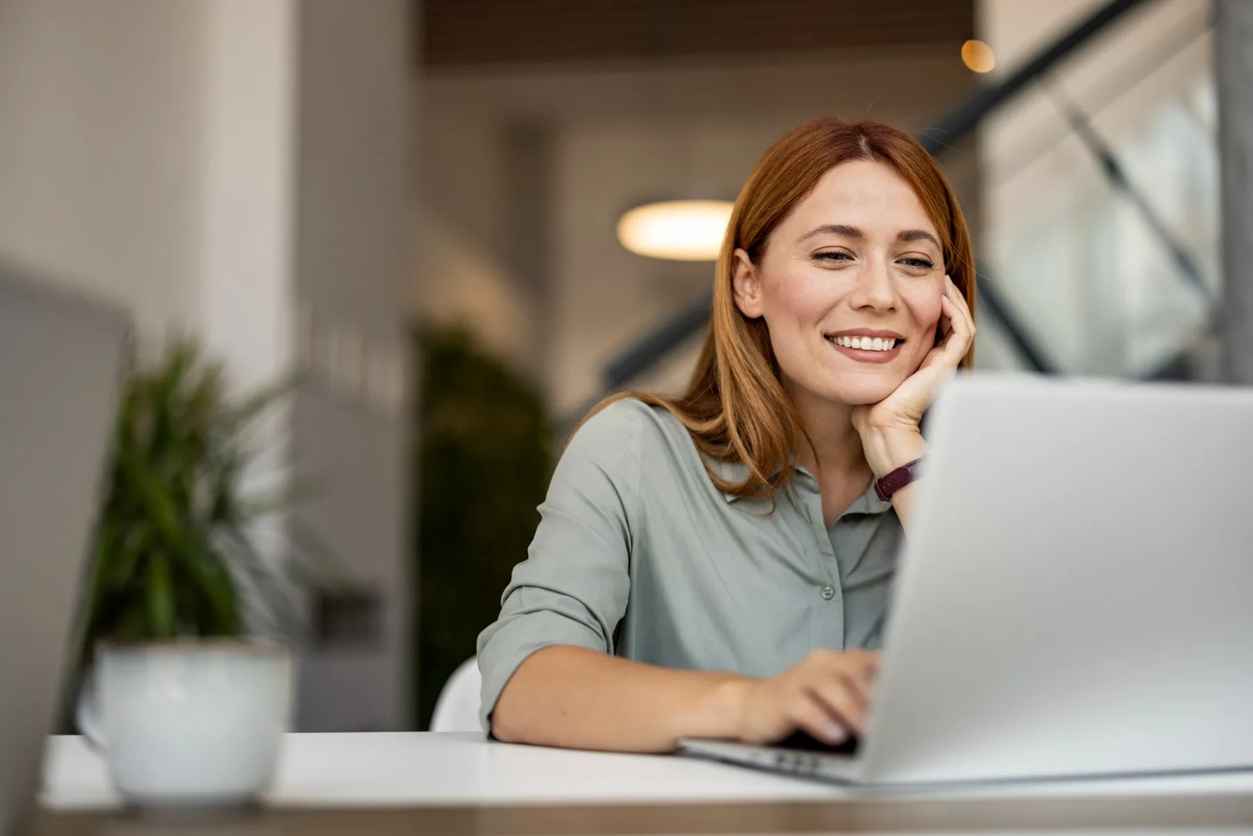 A smiling woman works on a laptop at an office desk, her hand resting on her cheek.