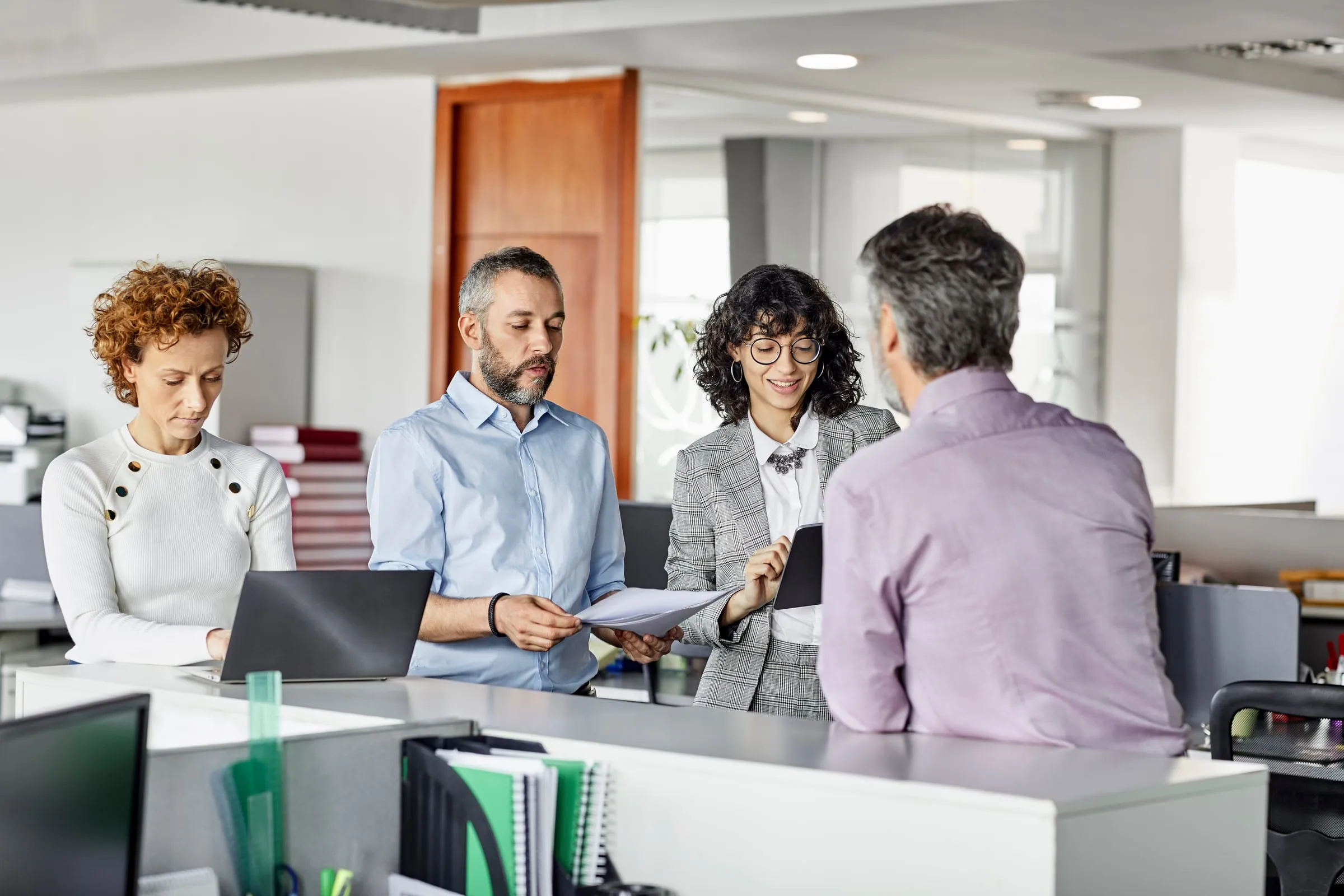 A person at an office counter attends to three colleagues reviewing documents, a tablet and a laptop.