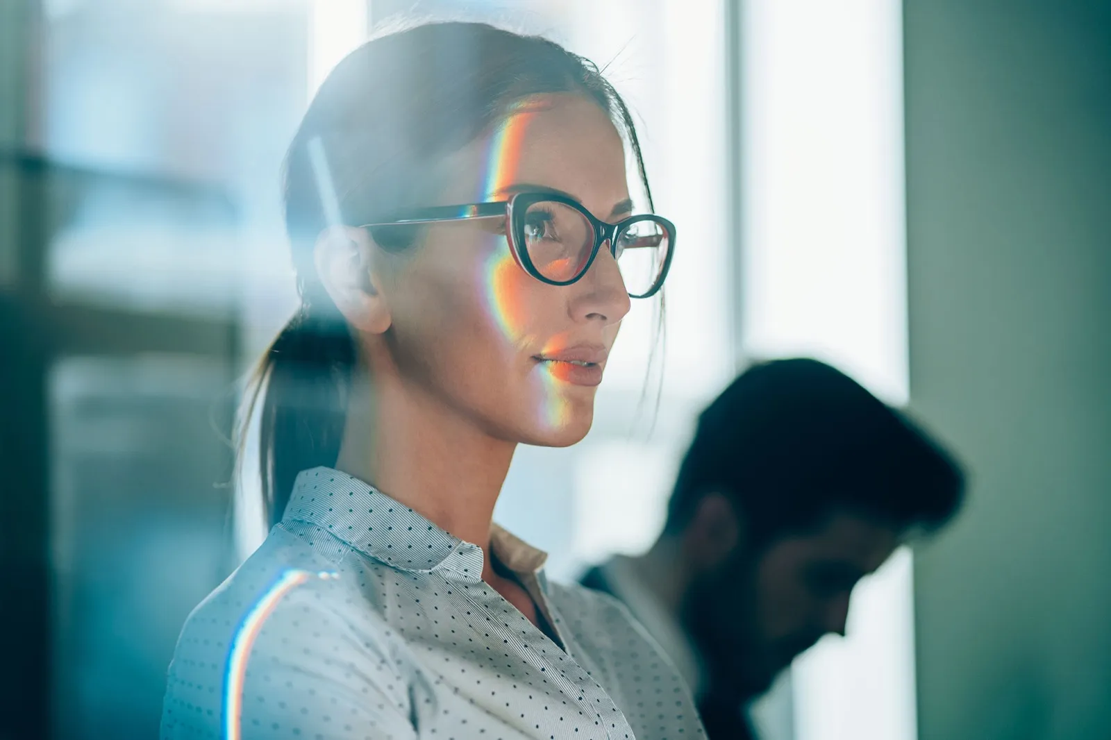 A woman wearing glasses looks to one side with an iridescent light reflection on her face and a blurred colleague behind her.