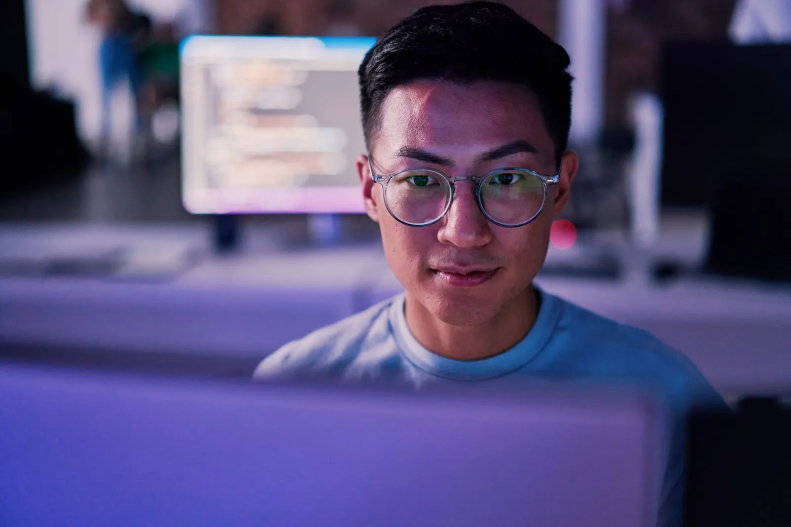 Un joven con gafas mira concentrado su pantalla en una oficina en penumbra iluminada en tonos violeta, con otro monitor con código al fondo.