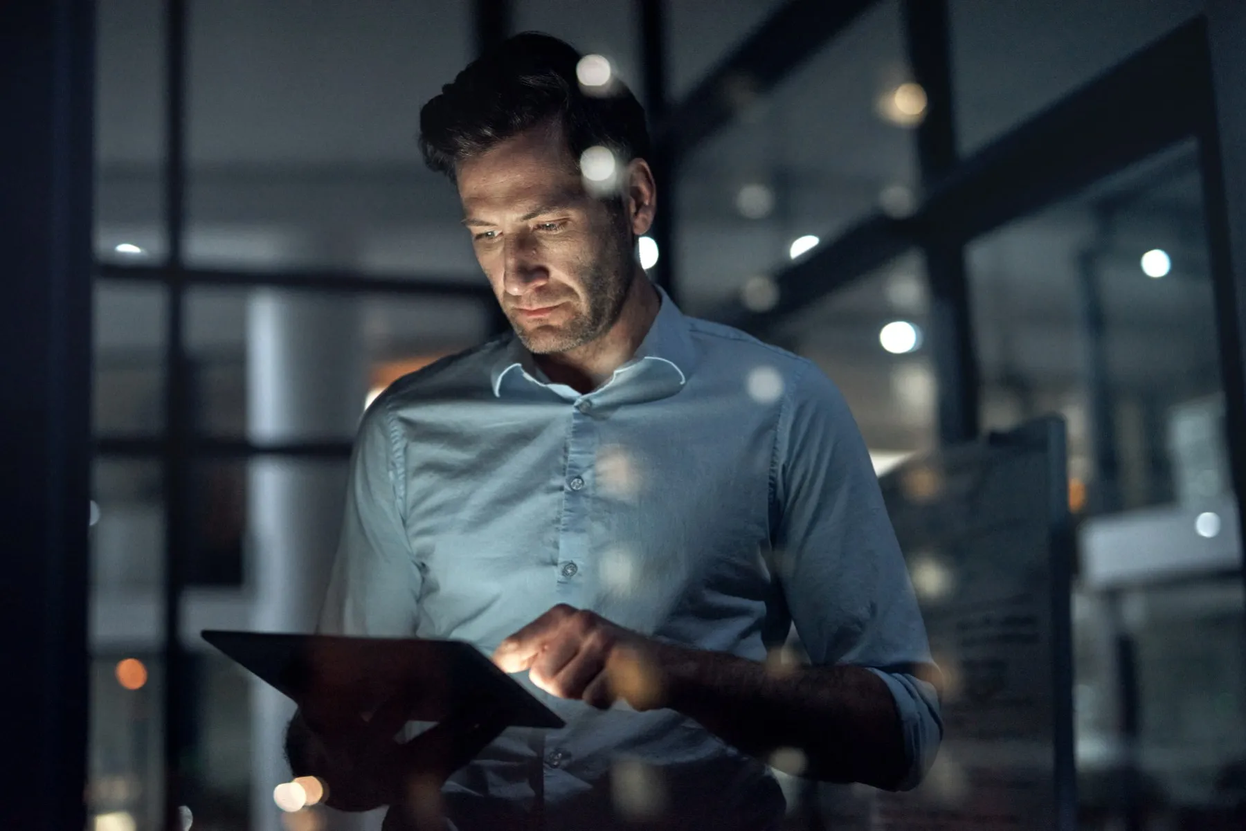 A person standing in a dimly lit office uses a tablet lit by its own screen.